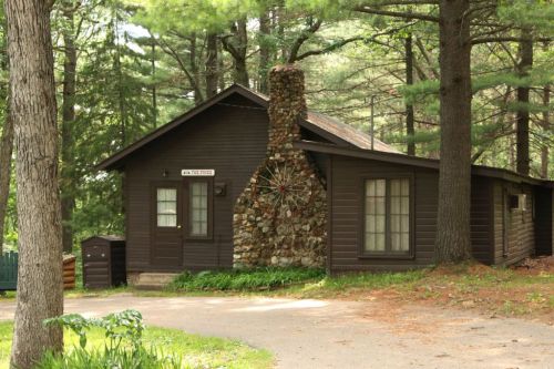 A rustic cabin with a stone chimney, surrounded by tall trees and a gravel path.