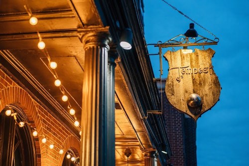A vintage sign for "Tempest" hangs outside a building, illuminated by warm string lights against a twilight sky.