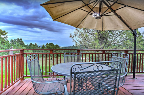 A patio with a round table and four chairs under a large umbrella, surrounded by trees and a cloudy sky.