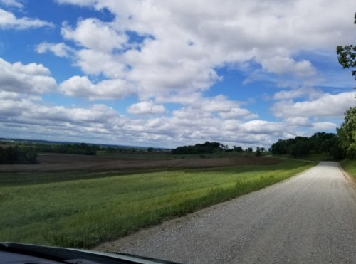 A gravel road winds through green fields under a partly cloudy blue sky. Trees line the horizon in the distance.