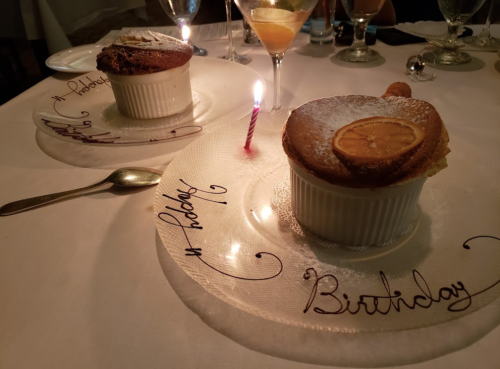 Two birthday desserts on elegant plates, one with a candle and orange slice, surrounded by glasses on a table.