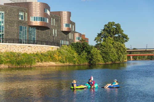 People relaxing on colorful floats in a river, with modern buildings and greenery in the background.