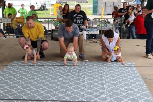 Babies crawl on a mat while adults cheer them on at a lively event under a tent.