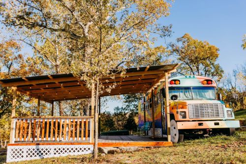 A colorful bus converted into a cozy outdoor seating area, surrounded by trees and autumn foliage.