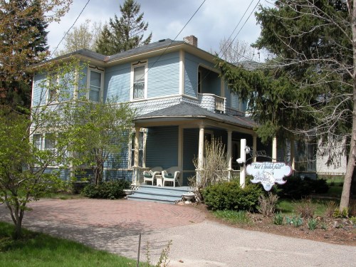 A blue Victorian-style house with a porch, surrounded by trees and a brick pathway.