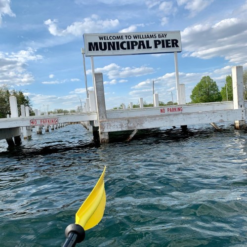 A yellow kayak paddle in the foreground, with a sign for "Municipal Pier" and a calm water scene in the background.