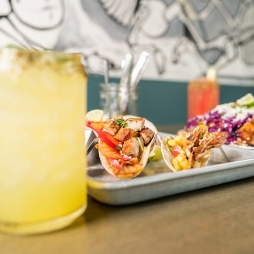 Two tacos filled with grilled meat and vegetables, served on a tray with a refreshing drink in the background.