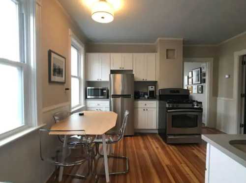 Bright kitchen with stainless steel appliances, a wooden table, and large windows, featuring light-colored cabinets and hardwood floors.