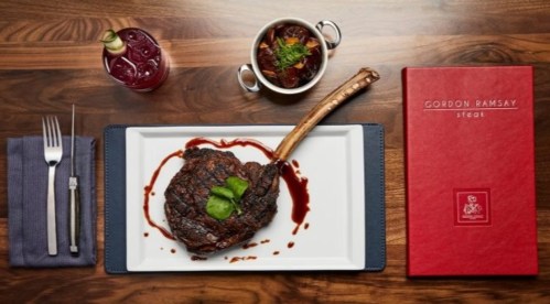 A gourmet steak on a white plate, accompanied by a side dish and a drink, with a menu on a wooden table.