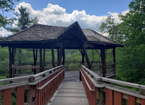 A wooden gazebo with a shingled roof, surrounded by lush green trees and a blue sky with clouds.