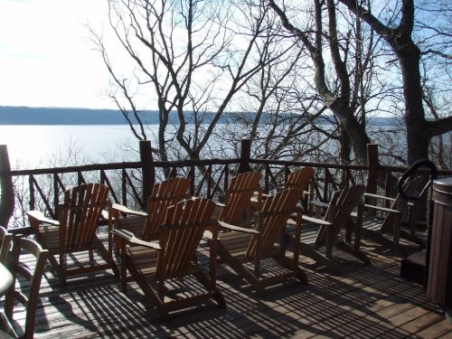 A wooden deck with several Adirondack chairs overlooking a calm lake, surrounded by bare trees and sunlight.
