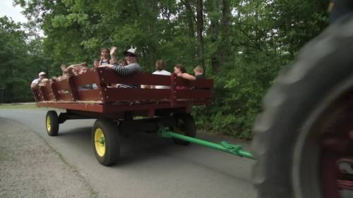A group of children and adults enjoy a ride on a wooden wagon pulled along a tree-lined path.