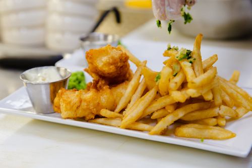A plate of golden fries with two pieces of fried fish and a small cup of dipping sauce, garnished with herbs.