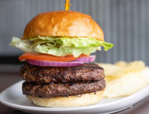 A stacked burger with two patties, lettuce, tomato, and onion, served with a side of potato chips on a white plate.
