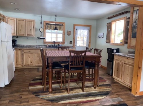 A cozy kitchen with wooden cabinets, a large table, and a rug, featuring natural light from windows.