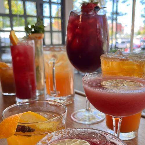 A variety of colorful cocktails on a table, featuring garnishes like fruit and herbs, in a bright setting.
