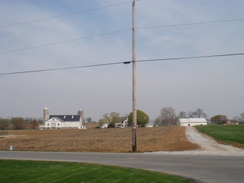 A rural landscape featuring a farm with silos, a dirt road, and a power pole under a clear sky.