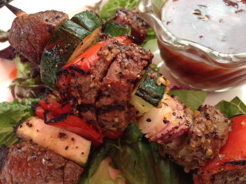 Grilled meat and vegetable skewers on a bed of greens, served with a side of dipping sauce.