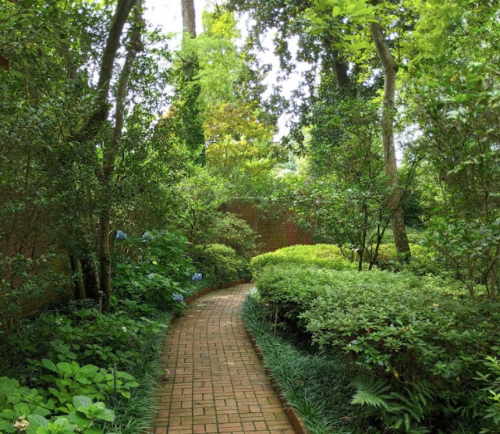 A winding brick path through a lush, green garden surrounded by trees and shrubs.