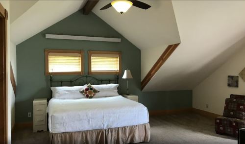 Cozy attic bedroom with a bed, decorative pillow, lamp, and sloped ceiling. Natural light from two windows.