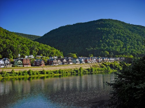 A serene river scene with a row of houses along the bank, surrounded by lush green hills under a clear blue sky.