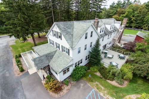 Aerial view of a large white house surrounded by trees, featuring a deck and outdoor seating areas.