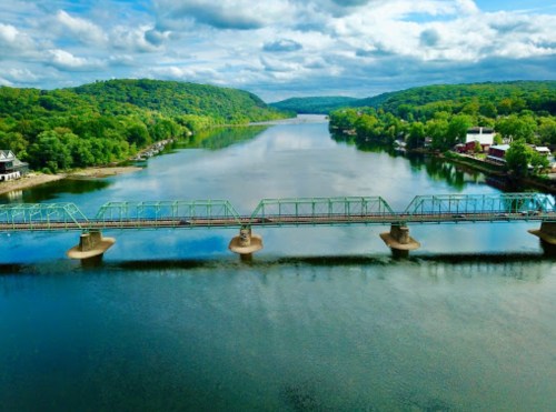 A scenic view of a green landscape with a bridge over a calm river, under a cloudy sky.