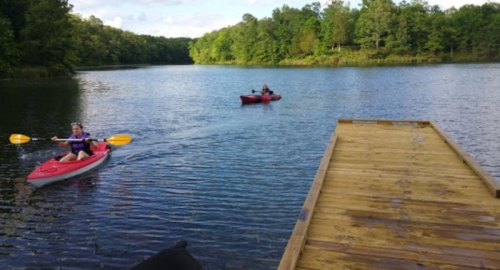 Two kayakers paddle on a calm lake, with a wooden dock in the foreground and lush greenery in the background.