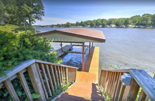 A wooden staircase leads down to a dock by a calm lake, surrounded by greenery and houses in the distance.