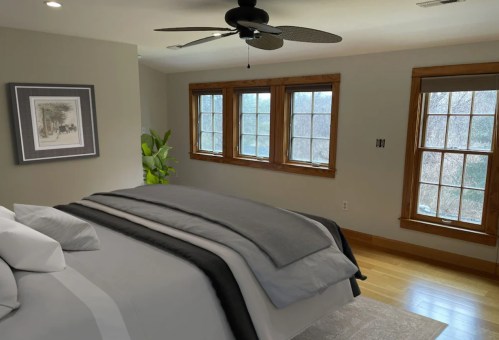 A cozy bedroom featuring a large bed, wooden windows, and a ceiling fan, with natural light and a plant in the corner.