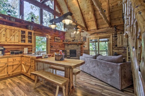 Cozy cabin interior featuring a wooden kitchen, dining table, and living area with a stone fireplace and large windows.