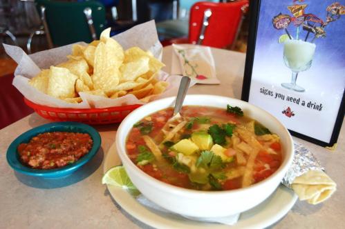 A bowl of soup with vegetables and tortilla strips, served with chips and salsa on a table with a drink sign.