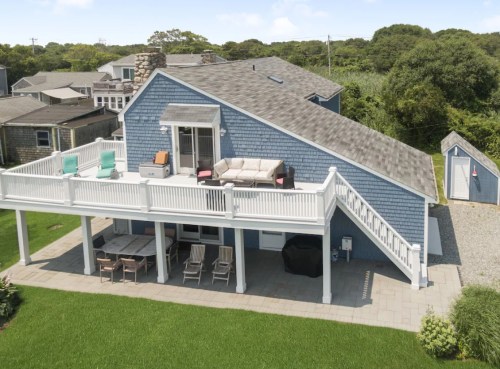A blue house with a large deck, outdoor furniture, and a grassy yard, surrounded by trees and other homes.