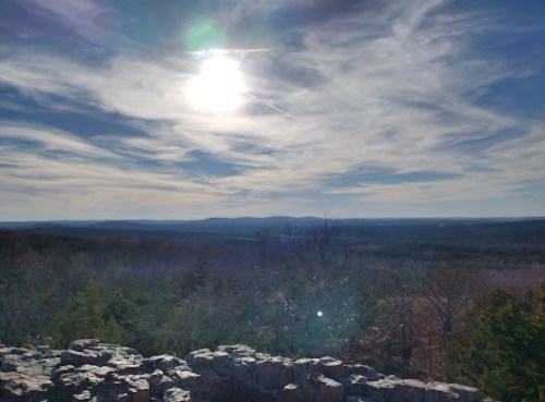 A scenic view of rolling hills under a bright sun and cloudy sky, with rocky foreground and trees in the distance.
