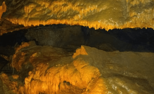 Stalactites and stalagmites in a dimly lit cave, showcasing orange mineral formations on the rocky surfaces.