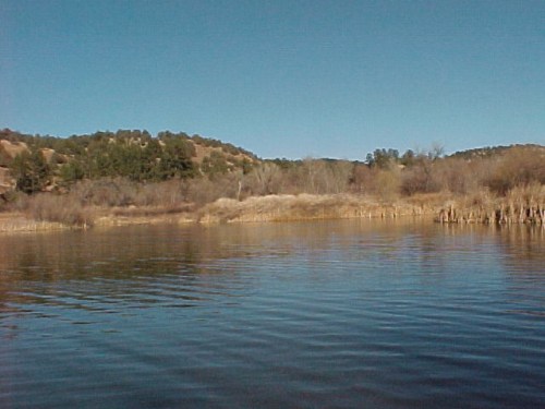 A serene lake surrounded by hills and trees under a clear blue sky, with gentle ripples on the water's surface.
