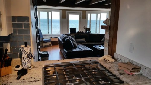 A view from a kitchen with a stove, looking into a living room with large windows overlooking the ocean.