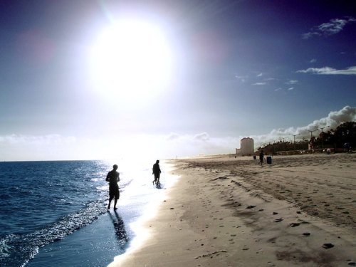Silhouettes of people walking along a sandy beach under a bright sun and clear blue sky.