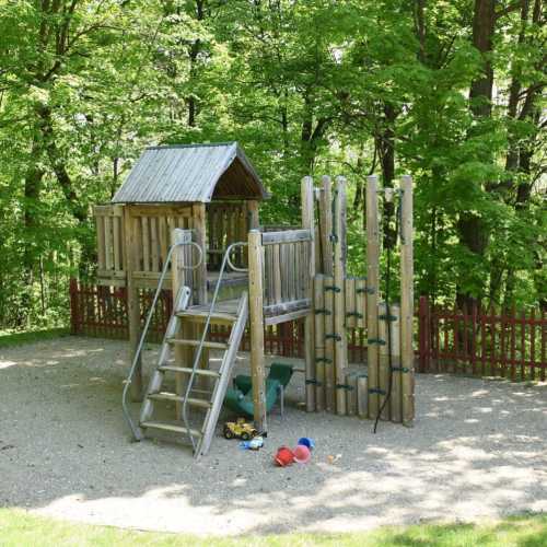 A wooden playground structure with a slide and climbing wall, surrounded by trees and a gravel play area.