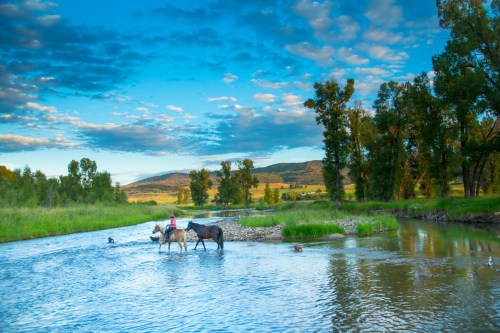 A serene river scene with two horses and riders crossing, surrounded by lush greenery and a blue sky with clouds.