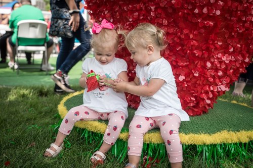Two young girls in matching outfits sit on a grassy area, playing with a toy strawberry near a large red heart decoration.