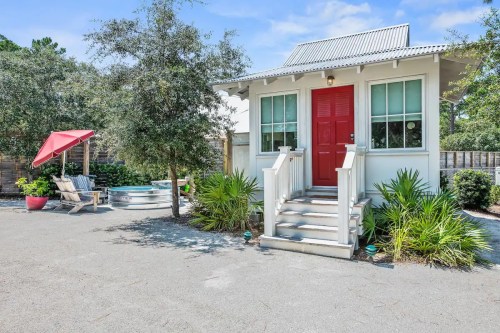 A small white cottage with a red door, surrounded by greenery and a patio with an umbrella and seating.