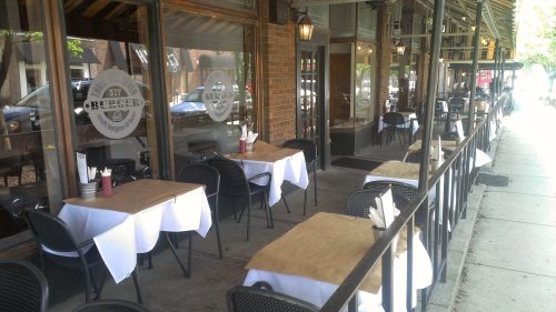 Outdoor dining area with empty tables and chairs, featuring white tablecloths and condiments on a sunny day.