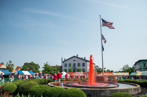 A vibrant fountain sprays orange water in a park, with flags flying and people gathered nearby under a clear blue sky.
