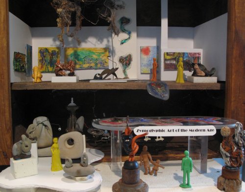 A display of eclectic art pieces, including sculptures and paintings, arranged on shelves and a table.