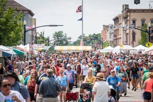 A bustling street fair with large crowds, tents, and vendors under a sunny sky. Flags and buildings line the street.