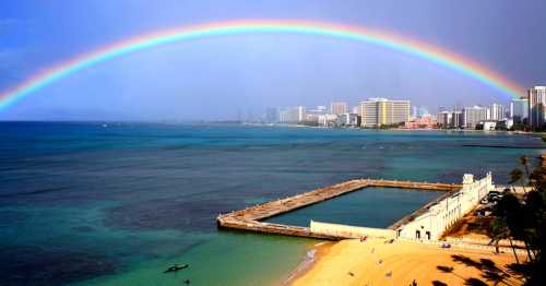 A vibrant rainbow arcs over a coastal city, with calm waters and sandy beach in the foreground.