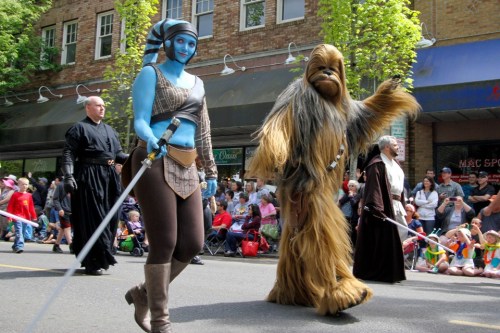 A parade scene featuring characters in costumes, including a blue-skinned figure and a large, furry creature, both with lightsabers.