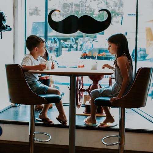 Two children sitting at a table in a café, enjoying ice cream and chatting, with a large mustache decoration in the window.