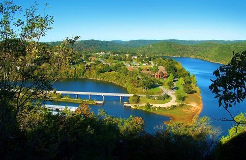 A scenic view of a river winding through green hills, with a bridge and houses visible along the shoreline.